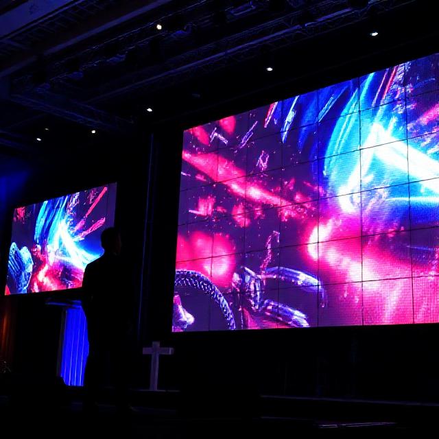 Large LED video wall displaying dynamic visuals at an event stage, clear pixels, presenter silhouette, realistic photo, high detail