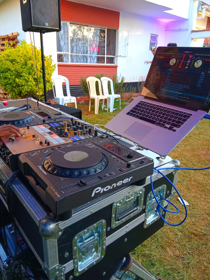 DJ performing at an event: modern DJ booth with turntables and mixer, headphones on, dancing crowd and colored lights in background, realistic photo, high detail